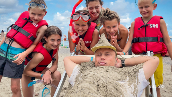 Jan Fitze im Liegestuhl, voll Sand, umringt von Kindern in Schwimmweste.