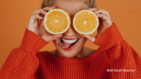 Frau in orangem Pulli hält sich zwei Orangenscheiben vor die Augen.